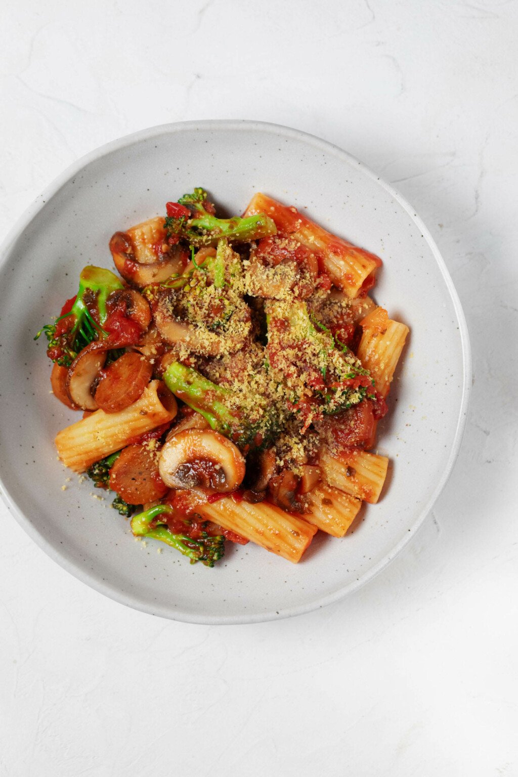 An overhead image of a white bowl, which is filled with a vegan pasta dish made with plant-based sausage, broccoli, and marinara sauce.