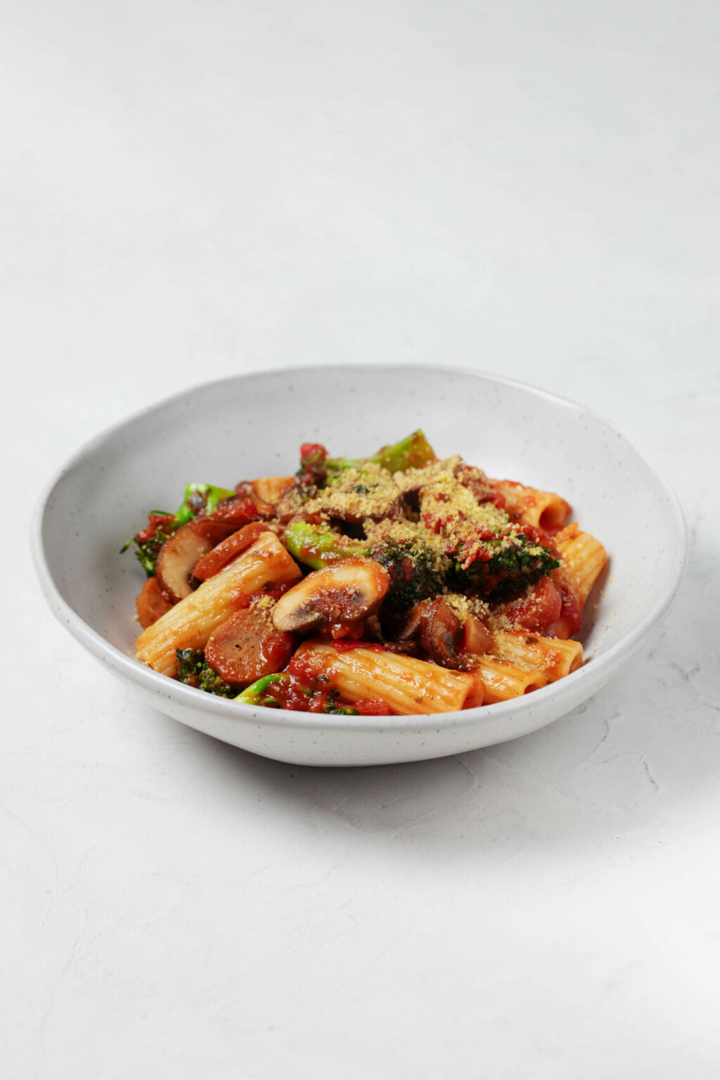 An angled image of a white bowl, which is filled with a vegan pasta dish made with plant-based sausage, broccoli, and marinara sauce.