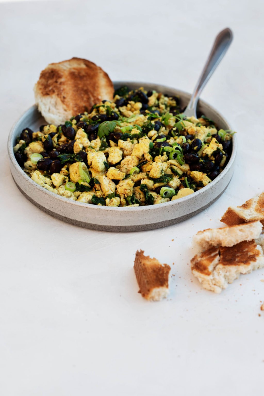 An angled photograph of a round plate, holding a vegan black bean tofu scramble with scallions and bread.