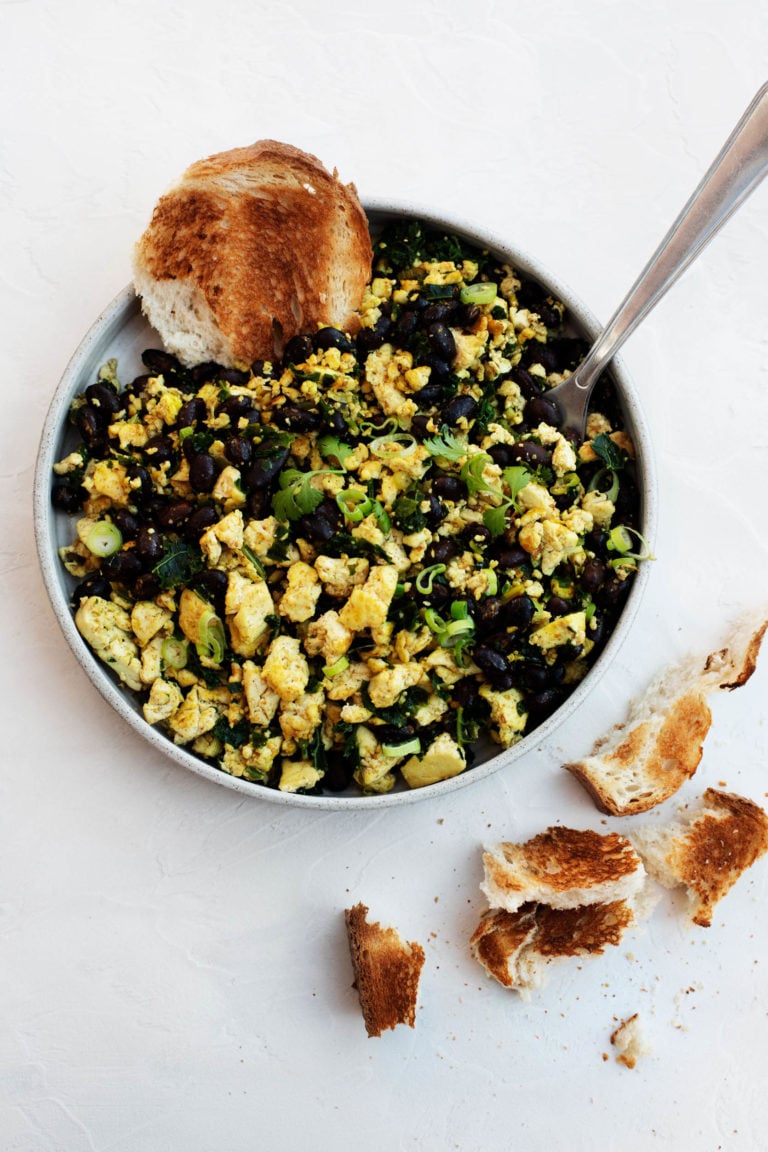 A round plate is filled with a black bean tofu scramble, made with chopped scallions and accompanied by small pieces of toast.
