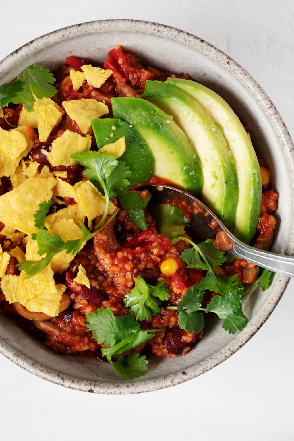 A close-up, overhead image of a bowl of hearty vegan mushroom bulgur bean chili. The red chili is topped with avocado slices and crumbled corn chips.