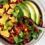 A close-up, overhead image of a bowl of hearty vegan mushroom bulgur bean chili. The red chili is topped with avocado slices and crumbled corn chips.