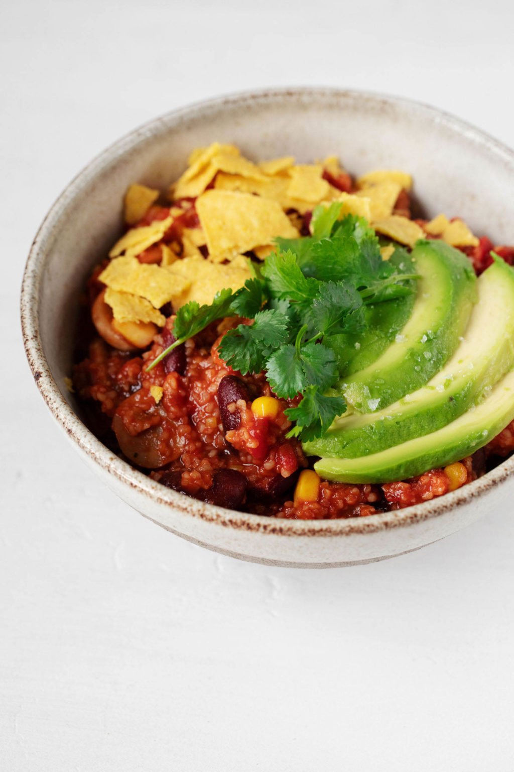 A white serving bowl rests on a white surface. It's holding the contents of a vegan bulgur bean chili, topped with avocado and fresh green herbs.