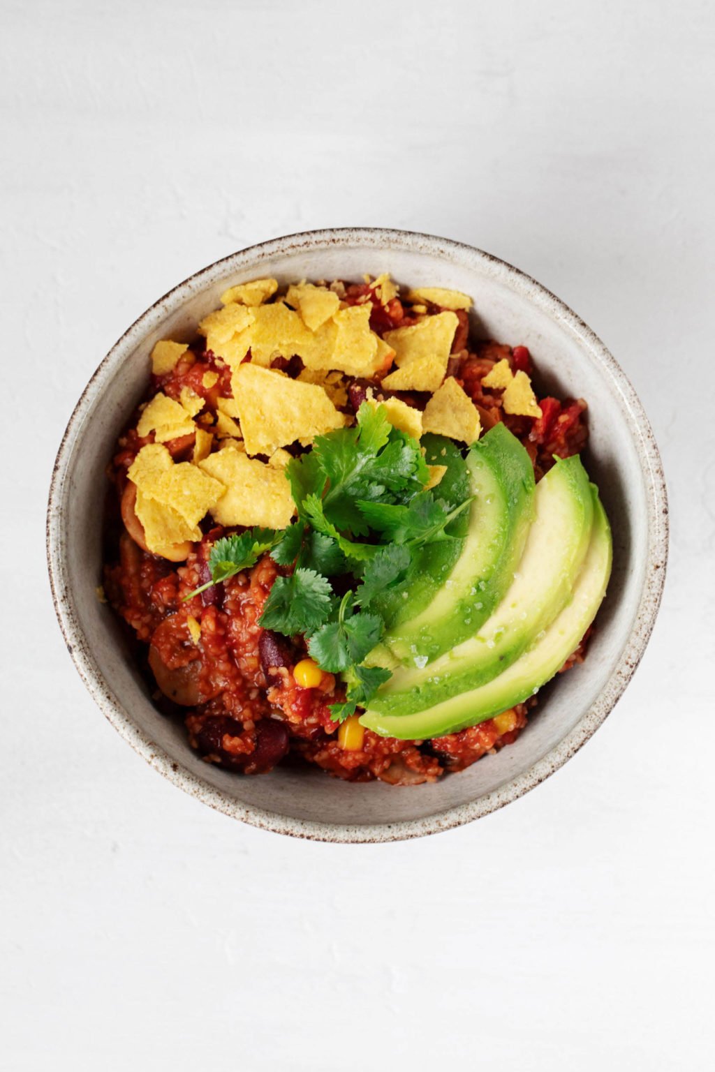 A white, round serving bowl holds a portion of a hearty vegan bulgur bean chili. It's topped with corn chips and avocado slices.