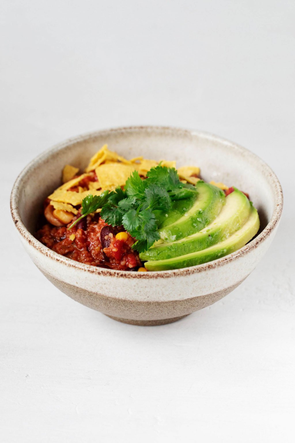An angled photograph of a cream colored and light brown bowl. It's filled with a bulgur and bean chili that has been garnished with avocado slices, corn chips, and green herbs.