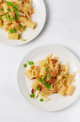 Two plates of caramelized cabbage onion pasta are resting on a white surface. Each has been lightly garnished with chopped parsley.