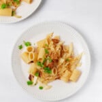 Two plates of a vegan cabbage and onion rigatoni dish are resting on a white surface. The pasta is garnished with flecks of green parsley.