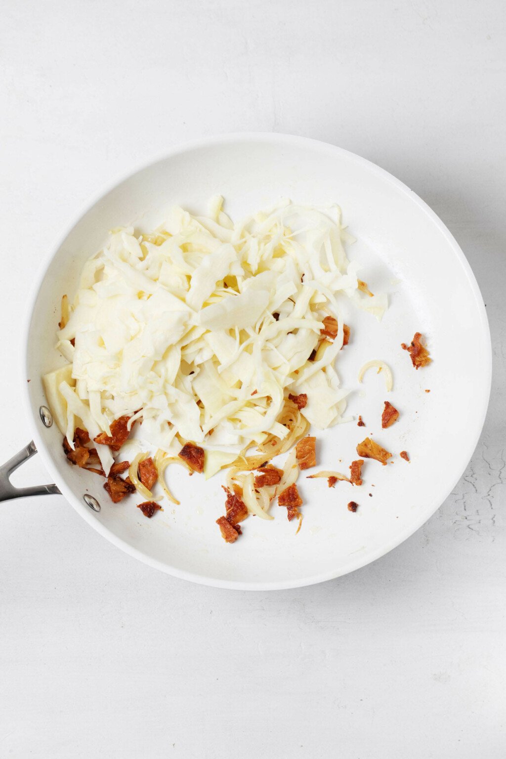 A white sauté pan has just been filled with sliced cabbage and plant-based bacon.