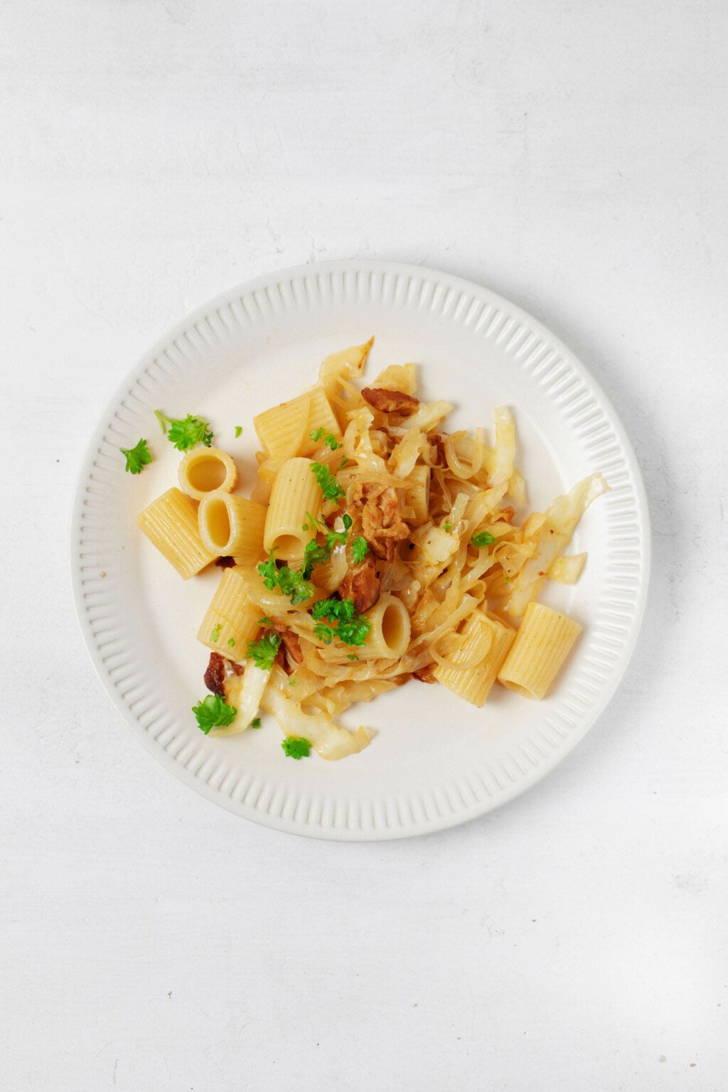 A plate of tube shaped pasta with caramelized cabbage and onion has been garnished with chopped green parsley.