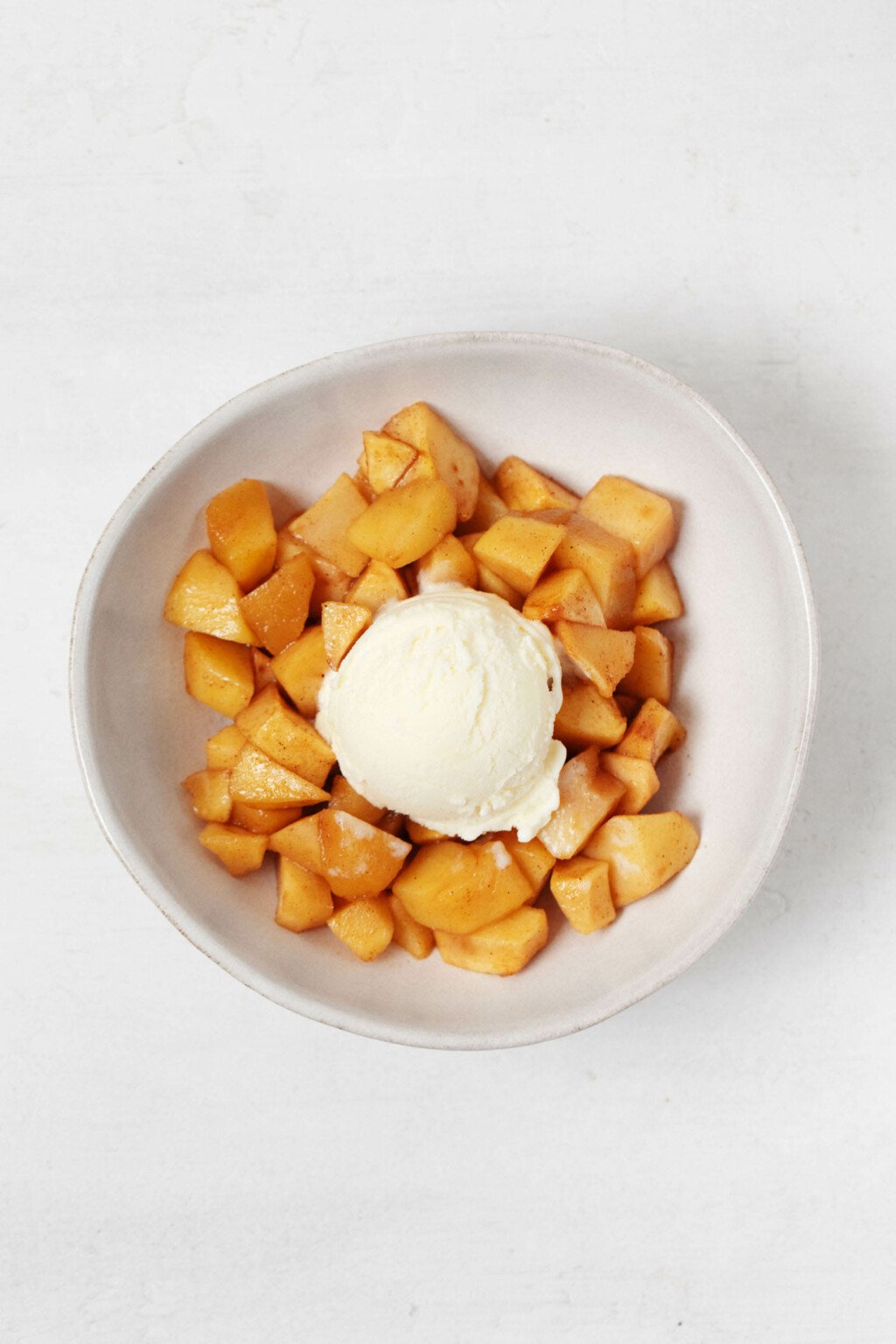 An image of a white bowl of baked apples, topped with a round scoop of vanilla ice cream.