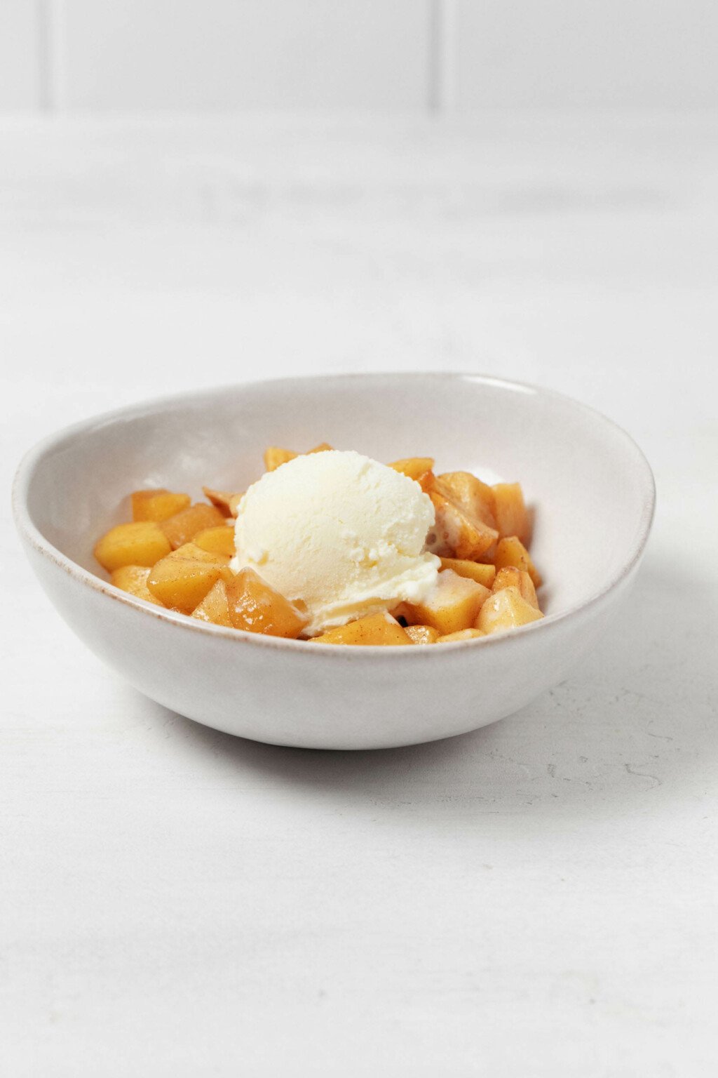 An angled image of a white ceramic bowl of baked apples, topped with a round scoop of vanilla ice cream.