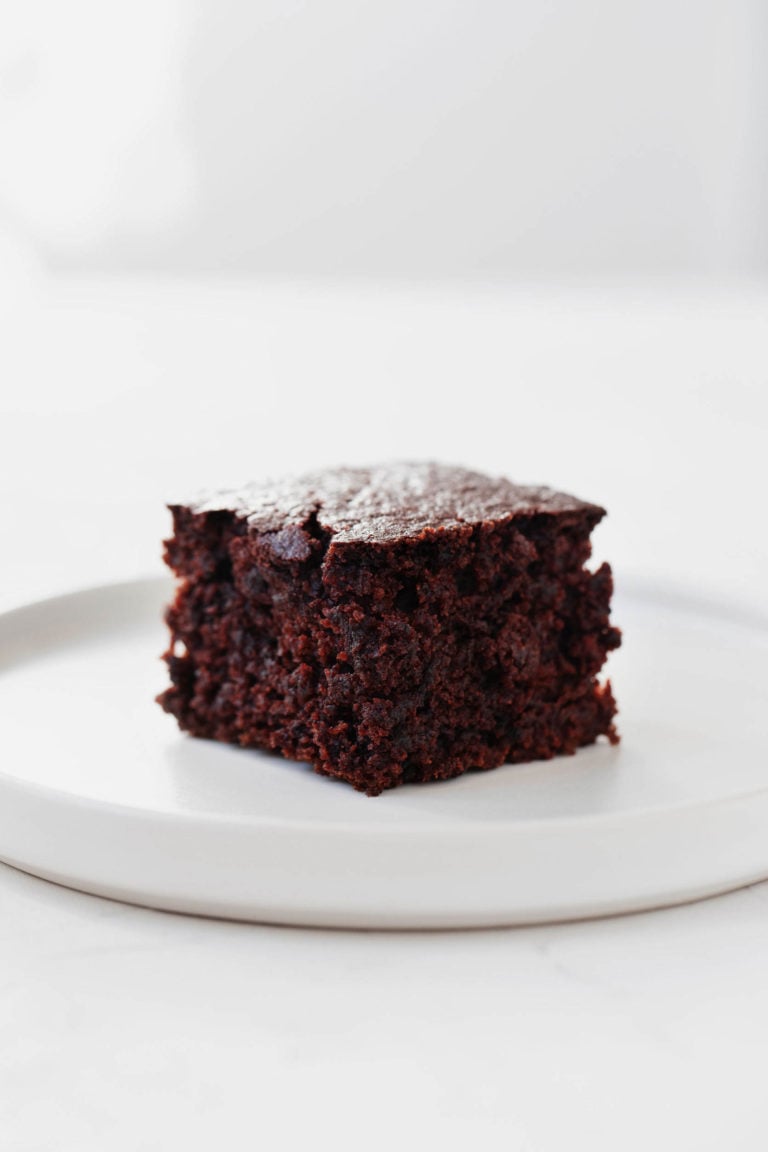 A round, white dessert plate holds a square slice of vegan chocolate beet cake. It rests on a white surface.