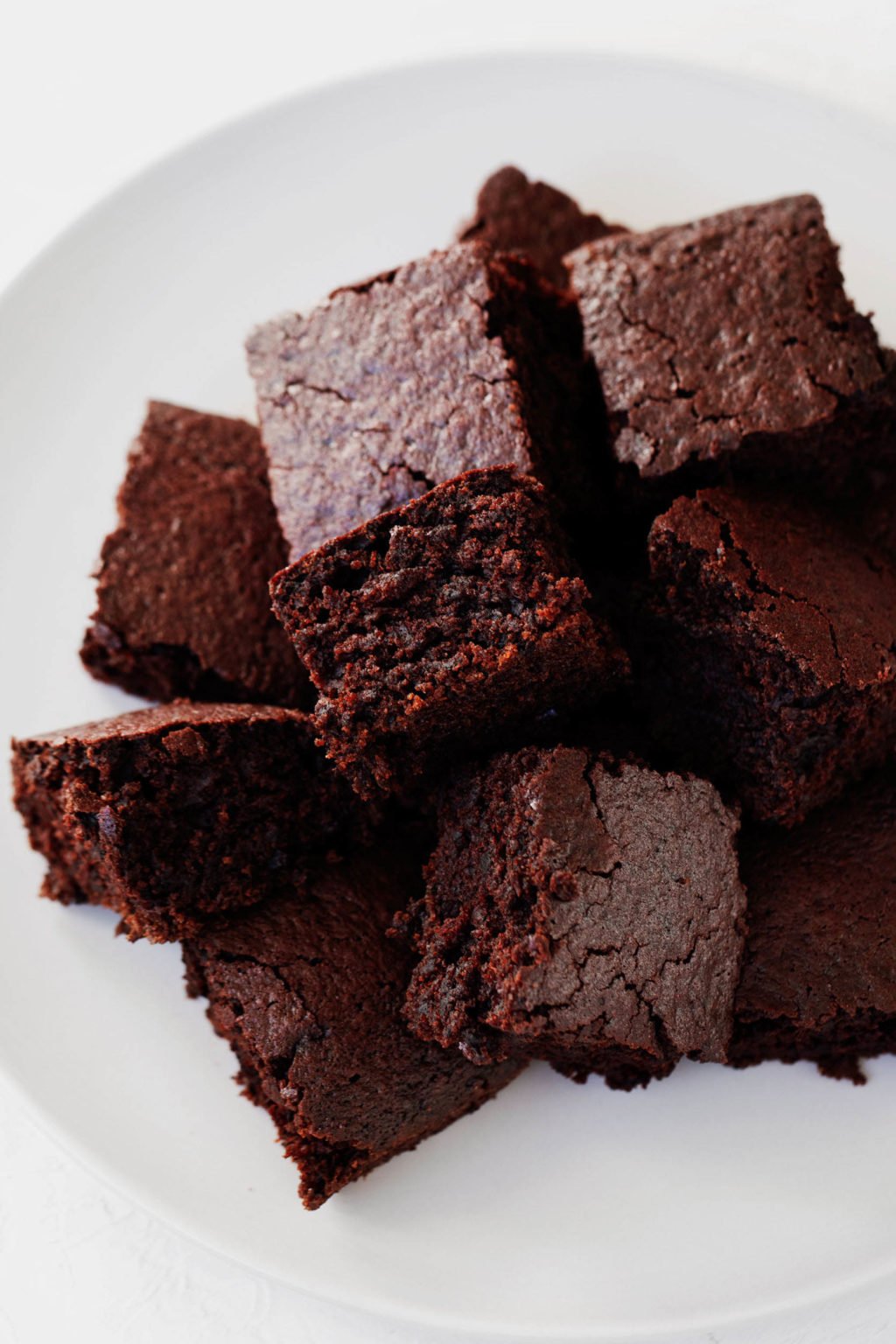 A small pile of chocolate cake slices rest on a white plate. The pieces are square and a deep brown color with red undertones.