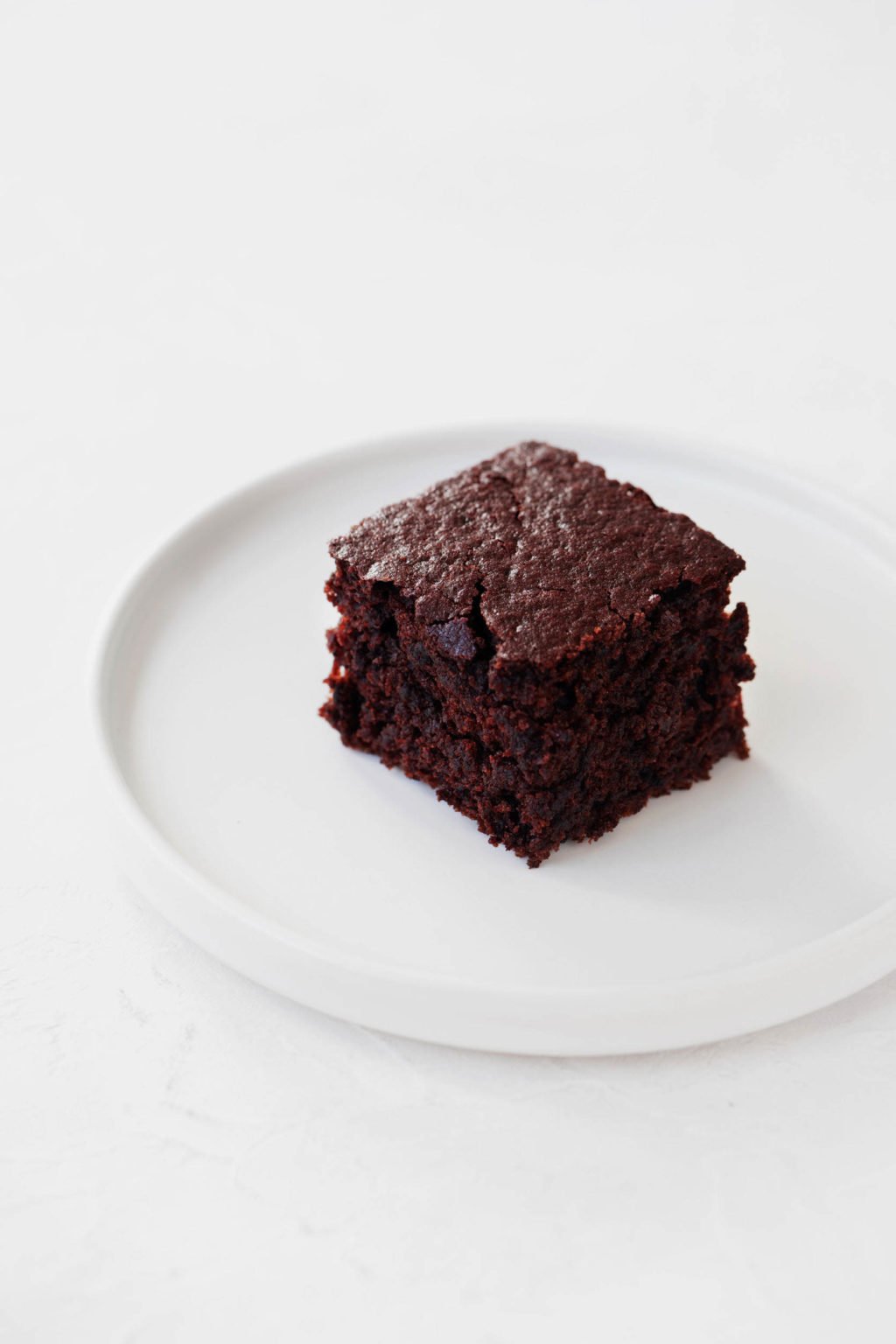 A small piece of plant-based, chocolate dessert rests on a white, round dessert plate.