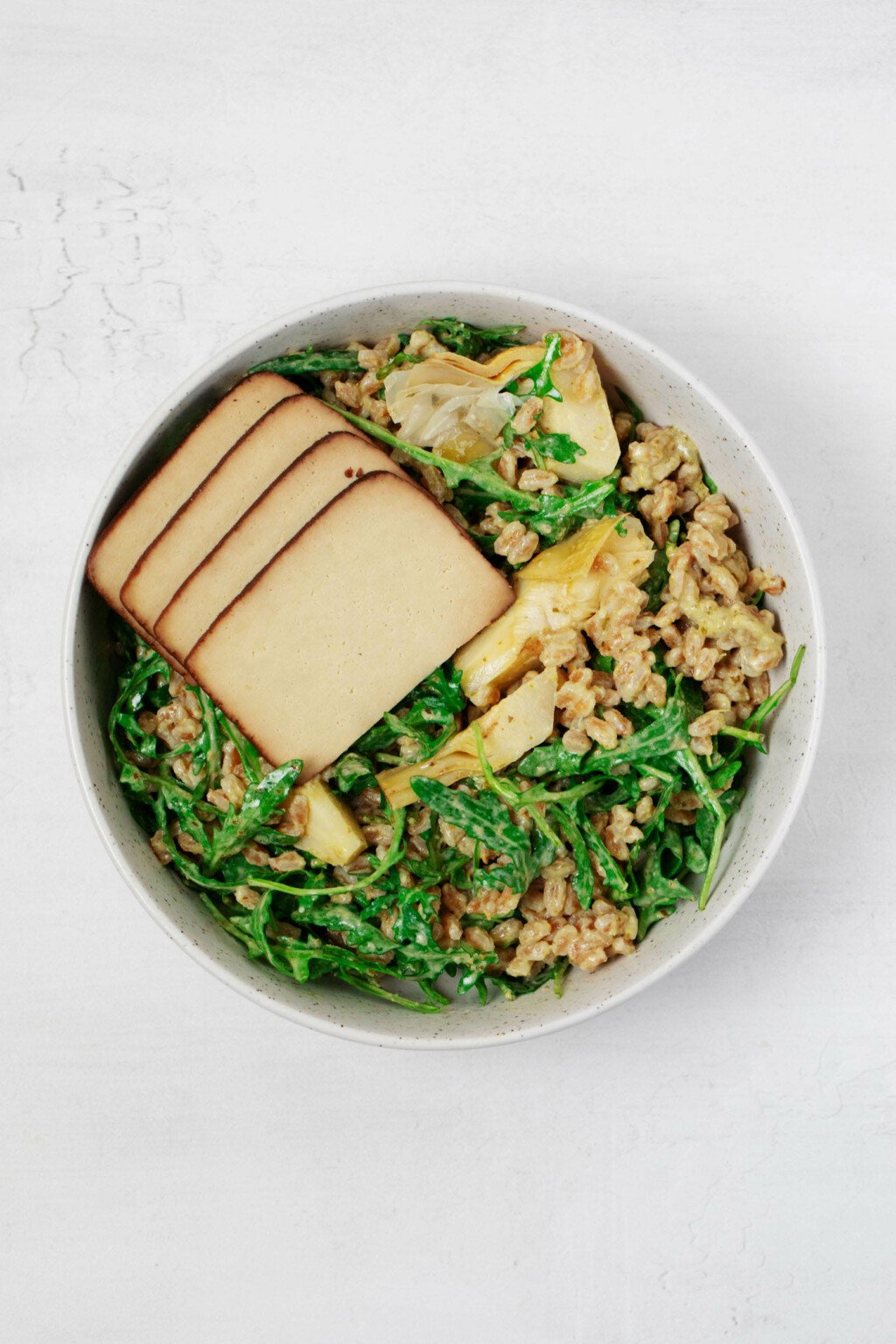 A round, white bowl is filled with whole grains, greens, and slabs of seasoned tofu.