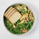A round, white bowl is filled with whole grains, greens, and slabs of seasoned tofu.
