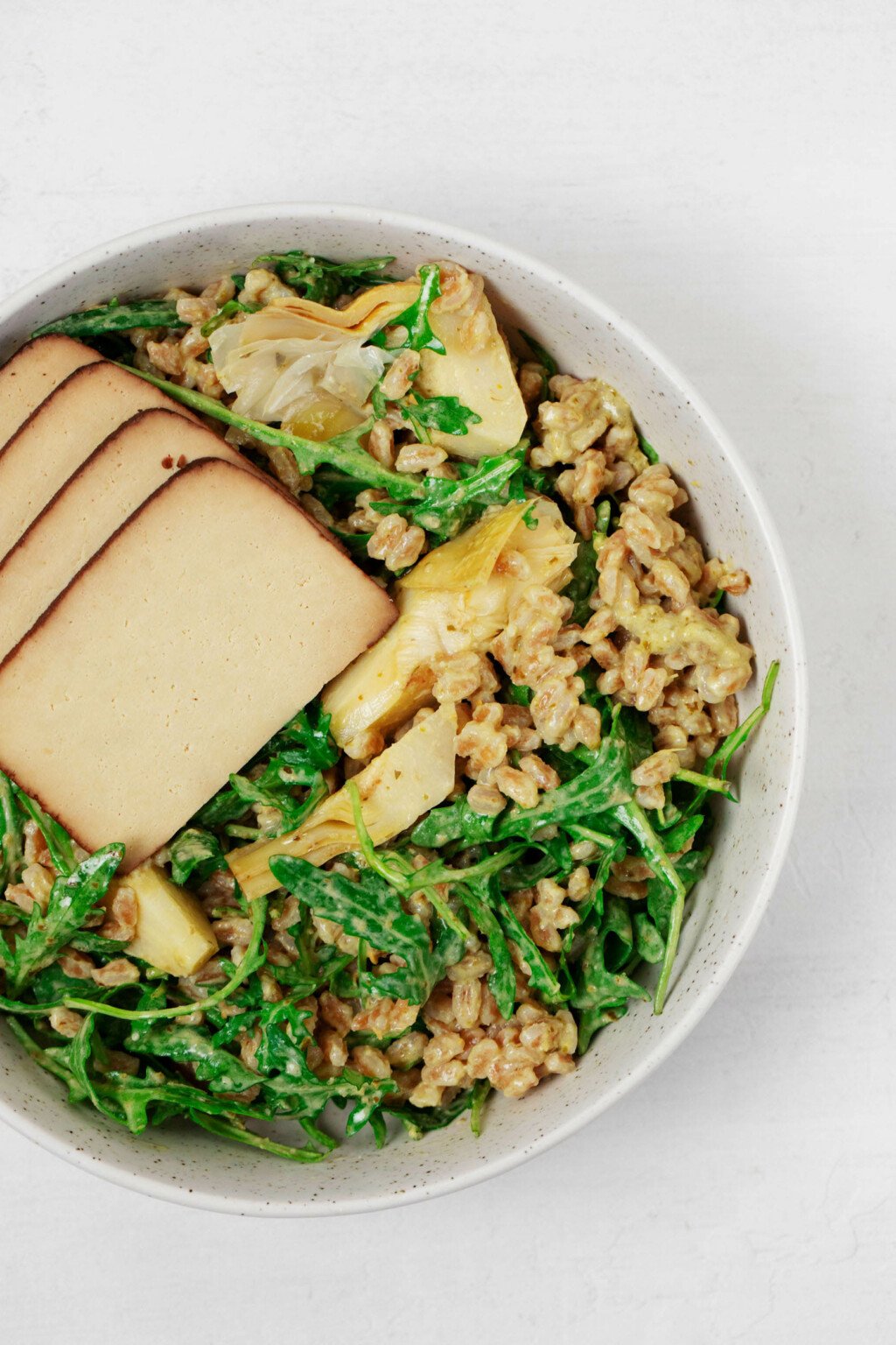 A close up image of a round, white bowl, which is filled with farro, kale, artichokes, and tofu. The farro is dressed with herby pesto.