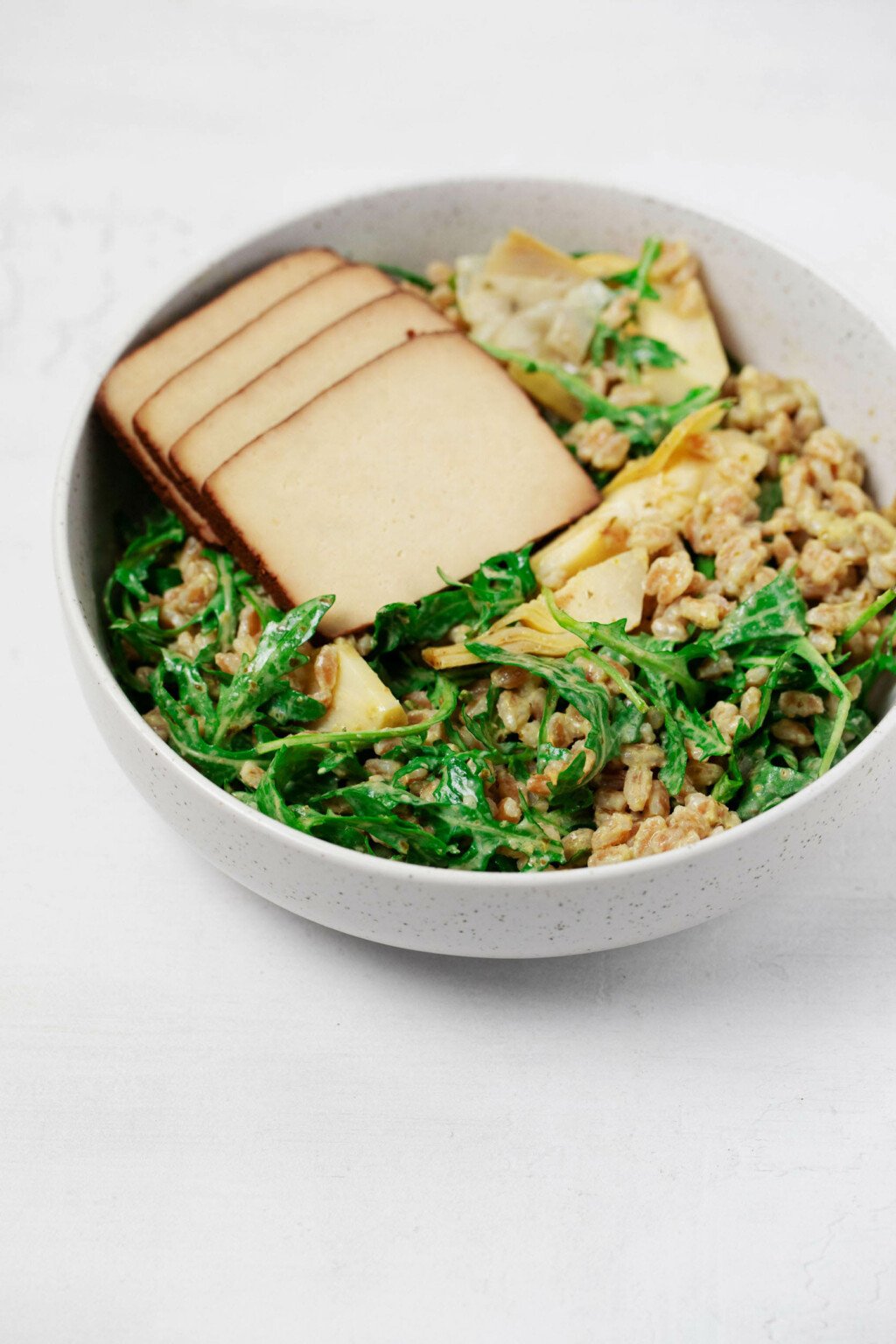 An angled, close up image of a round, white bowl, which is filled with farro, kale, artichokes, and tofu. The farro is dressed with herby pesto.