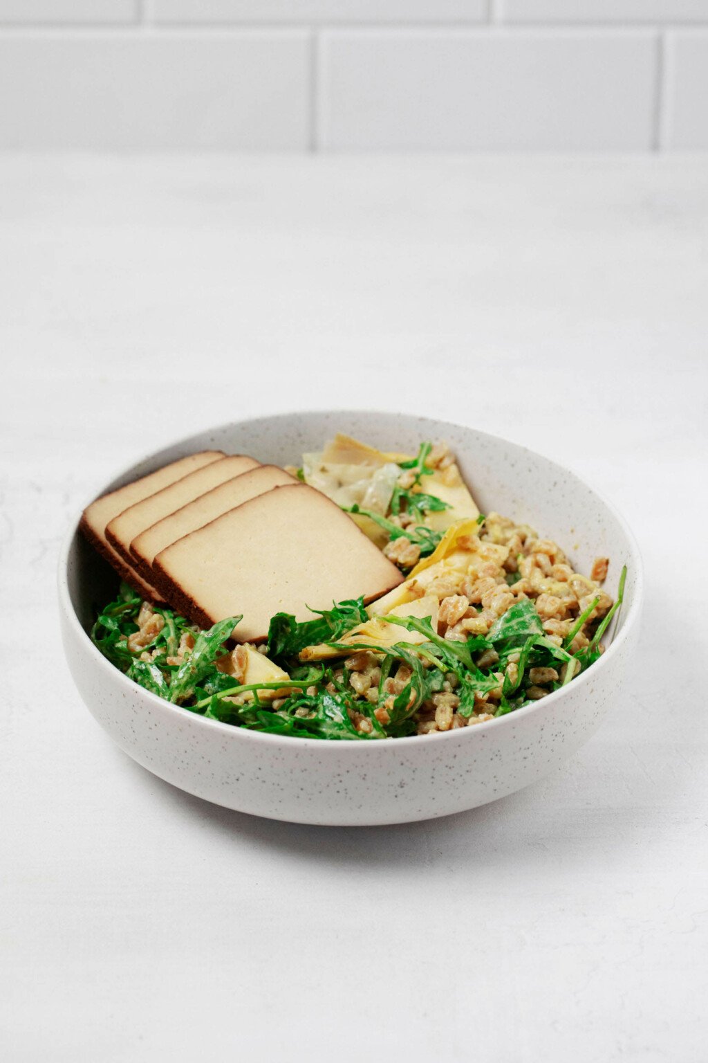 A large, white bowl is resting on a white surface against a white tiled wall. It holds the ingredients for a plant-based grain salad with kale.