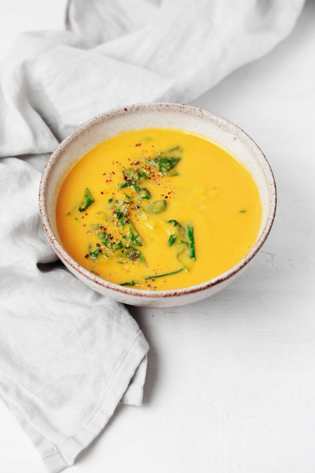 A gray, ceramic bowl of pureed chickpea soup is resting on a white surface. A gray, crumpled napkin rests nearby.