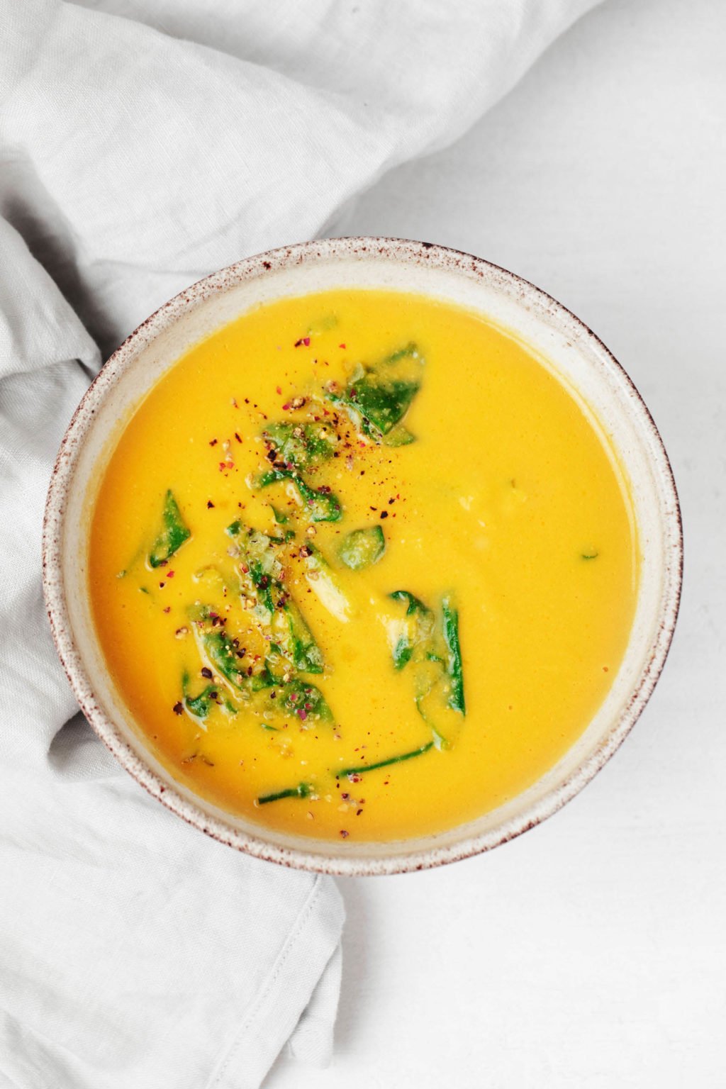 A round, gray and white ceramic bowl is filled with a roasted garlic and pureed chickpea soup with wilted greens.