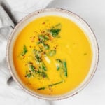 A round, gray and white ceramic bowl is filled with a roasted garlic and pureed chickpea soup with wilted greens.