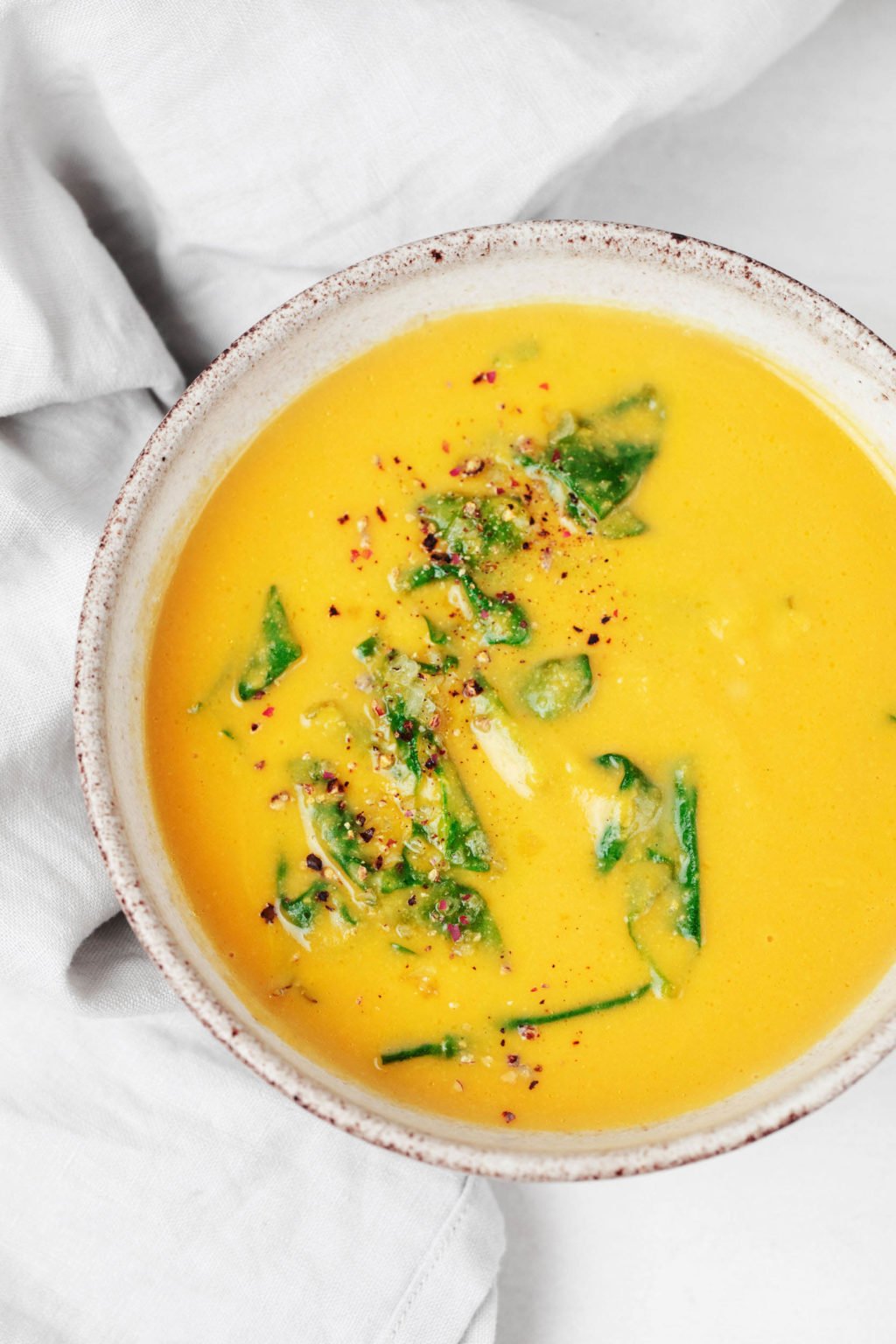 A bowl of pureed chickpea soup has been topped with wilted greens and pepper flakes. It rests on a white surface with a gray napkin nearby.