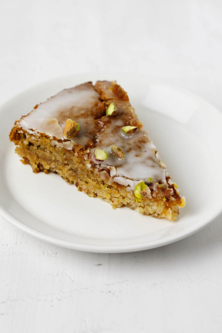 A slice of glazed vegan lemon pistachio cake rests on a small, round white dessert plate.
