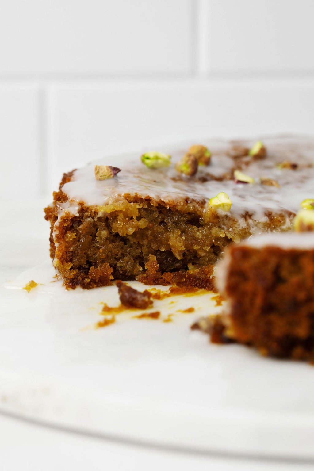 A glazed cake has been topped with chopped nuts. A slice has been removed. The cake is pictured against a white backdrop.