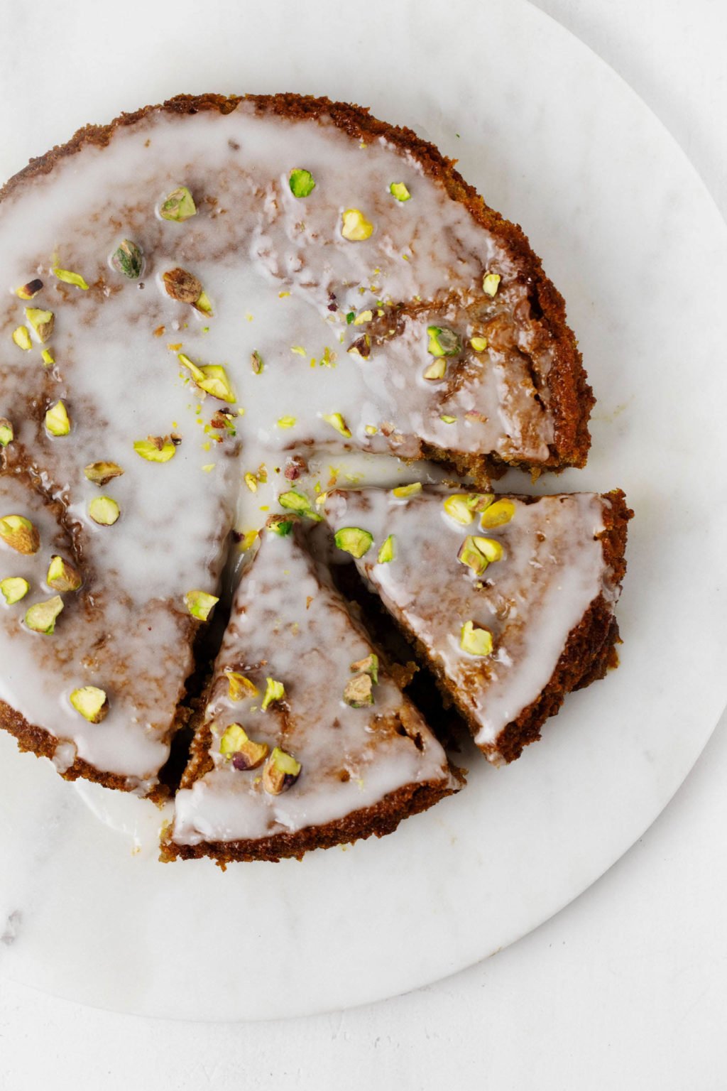 An overhead image of a vegan lemon pistachio cake. It has been cut into triangular slices and topped with a white, lemon glaze.