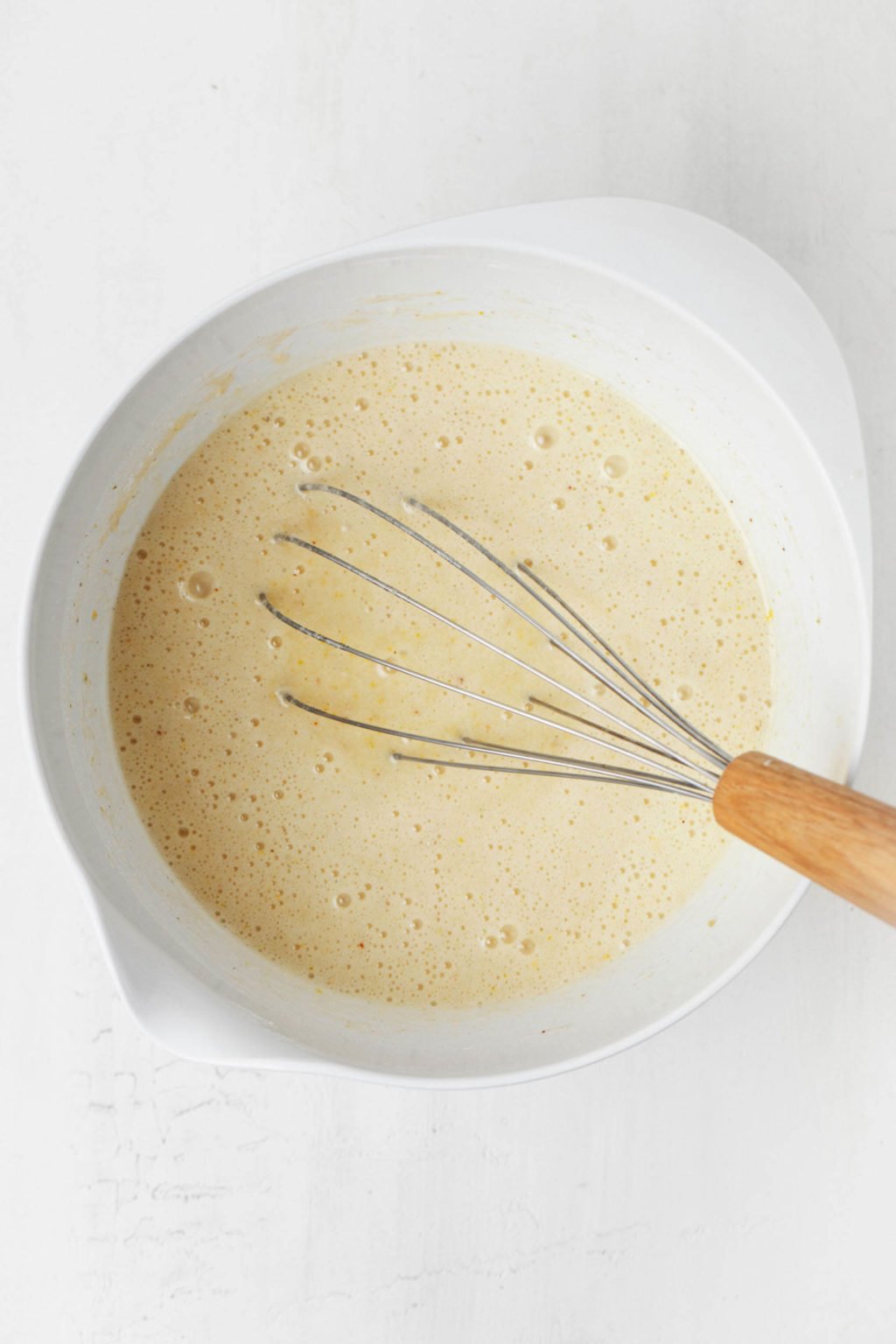 Cake batter is pictured in a white mixing bowl, along with a silver whisk with a wooden handle..