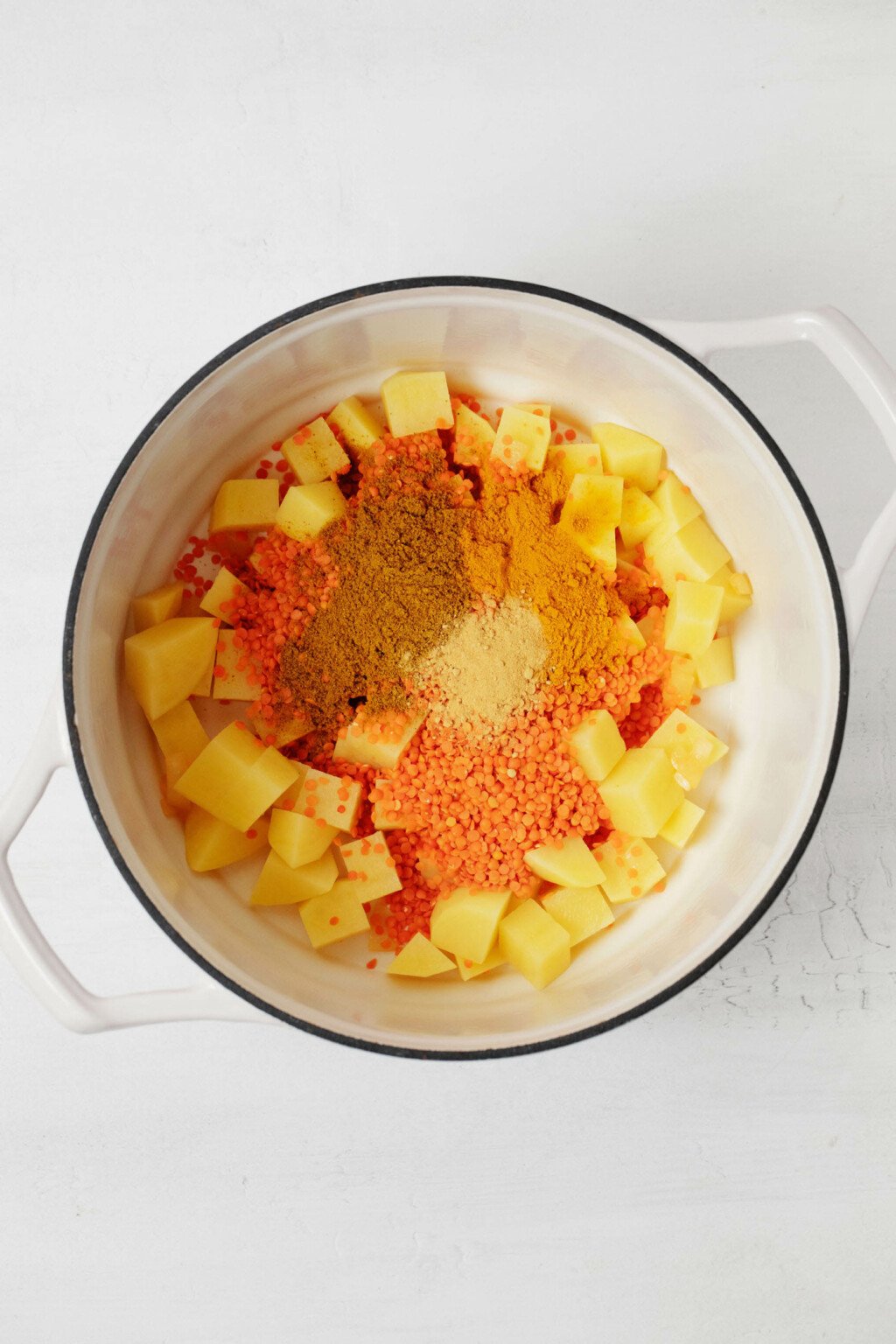 A round, white pot rests on a white surface. It's filled with potato cubes and spices.