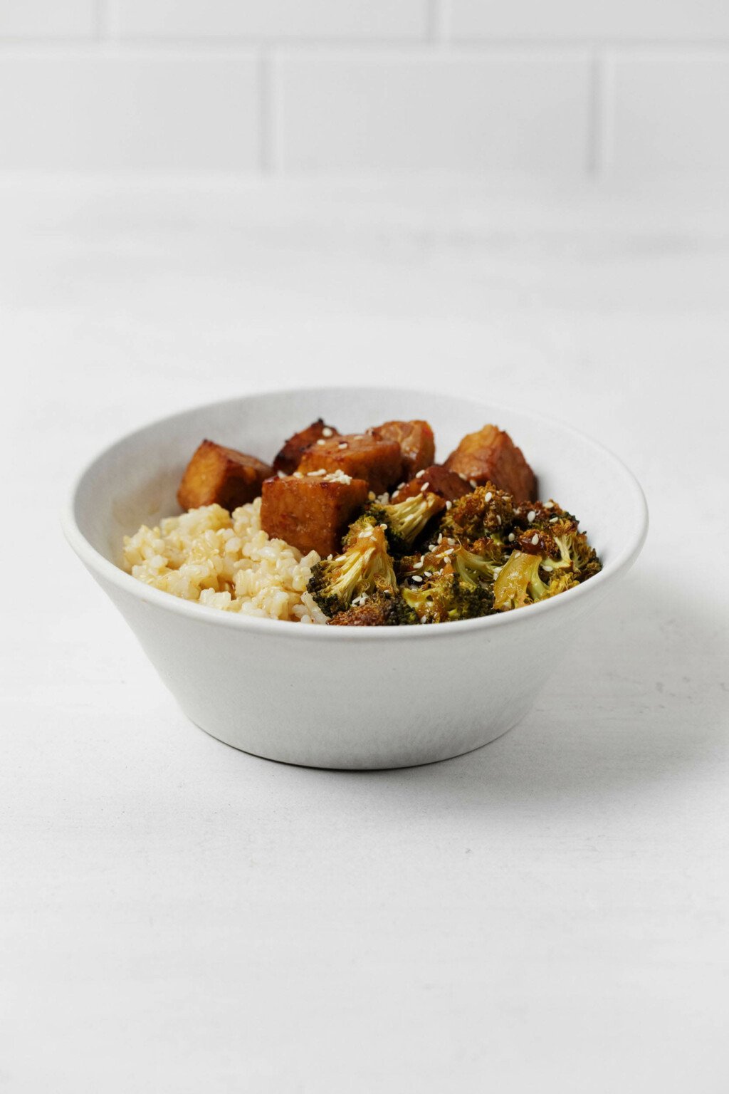 A wide, white ceramic bowl holds cubes of tempeh, broccoli, and brown rice.