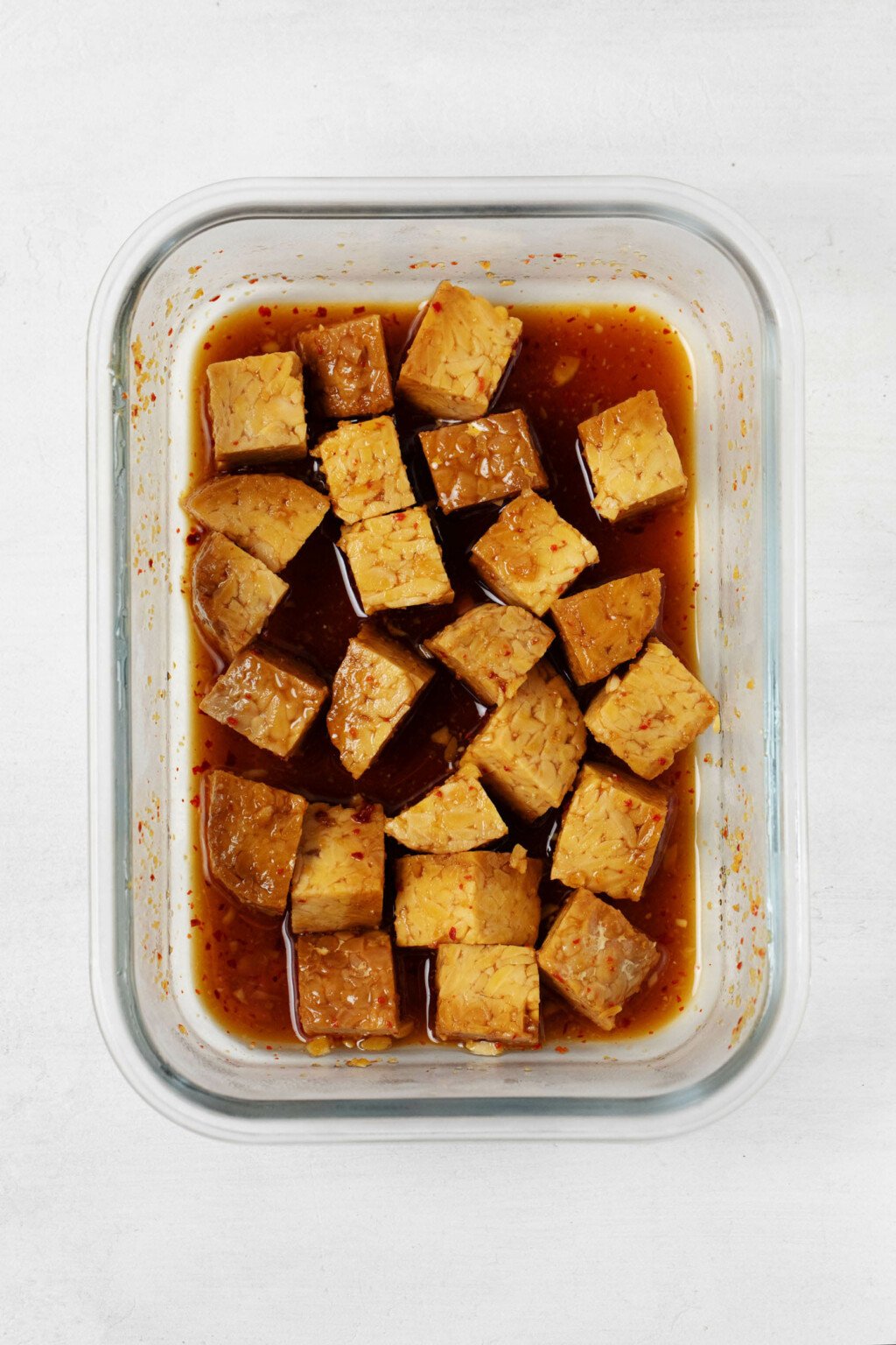 A clear, rectangular storage dish holds pieces of tempeh mixed with a dark colored sauce.