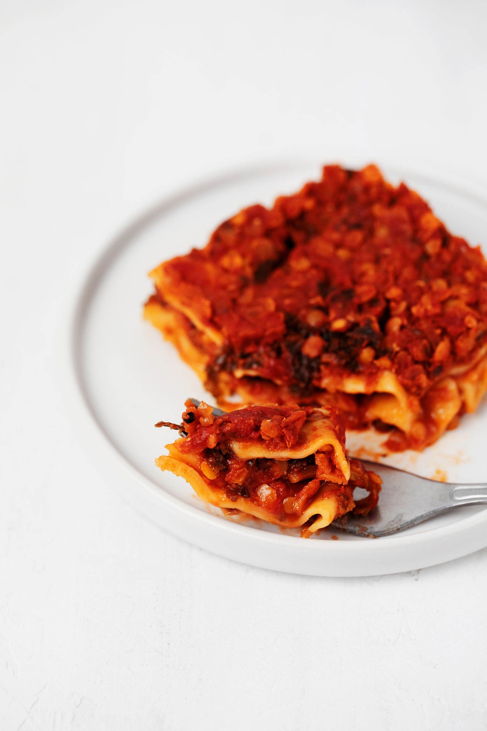 A slice of vegan lasagna with lentils and kale has just been cut into with a fork. It rests on a small, white plate.