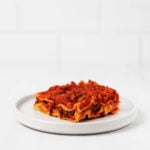 A slice of kale lasagna, topped with red sauce, is served on a small, round, white plate. It rests on a white surface.