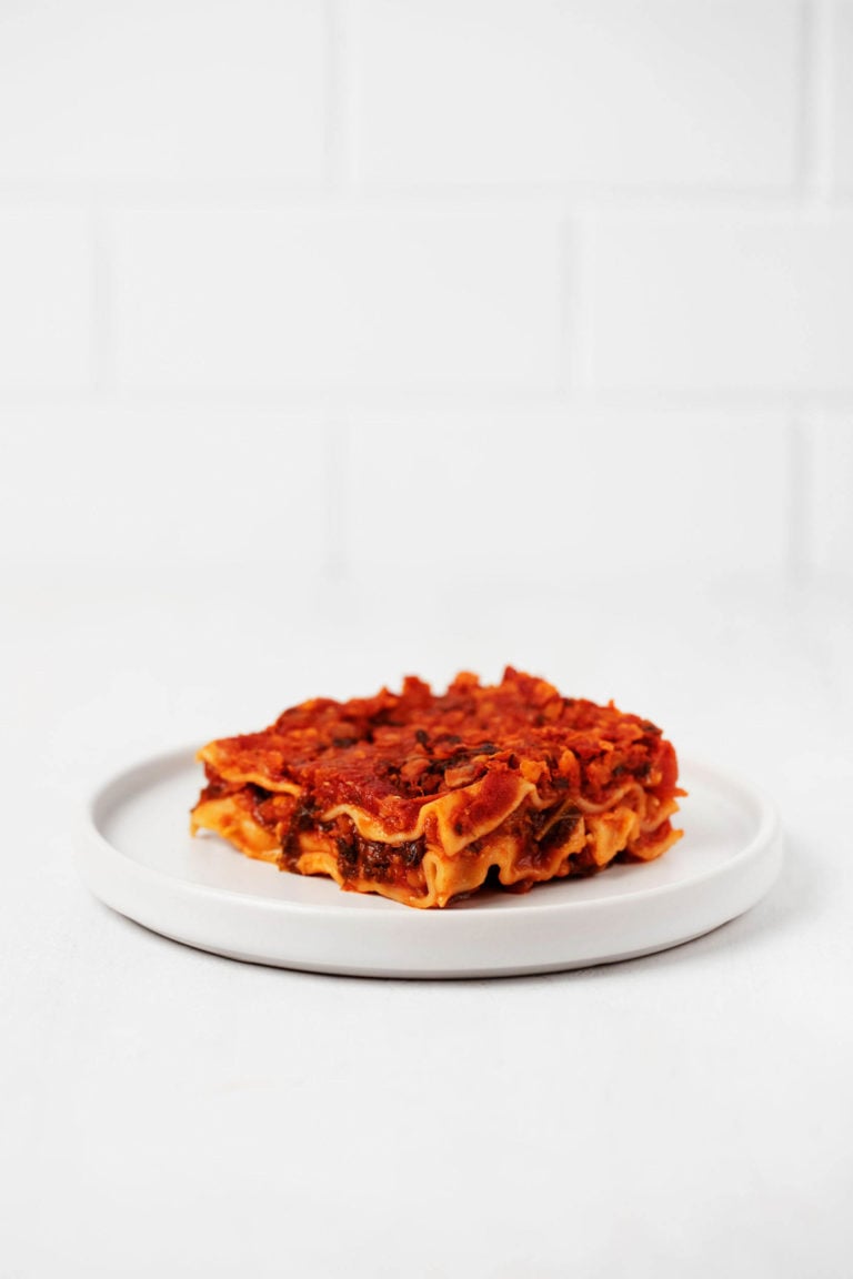 A slice of kale lasagna, topped with red sauce, is served on a small, round, white plate. It rests on a white surface.
