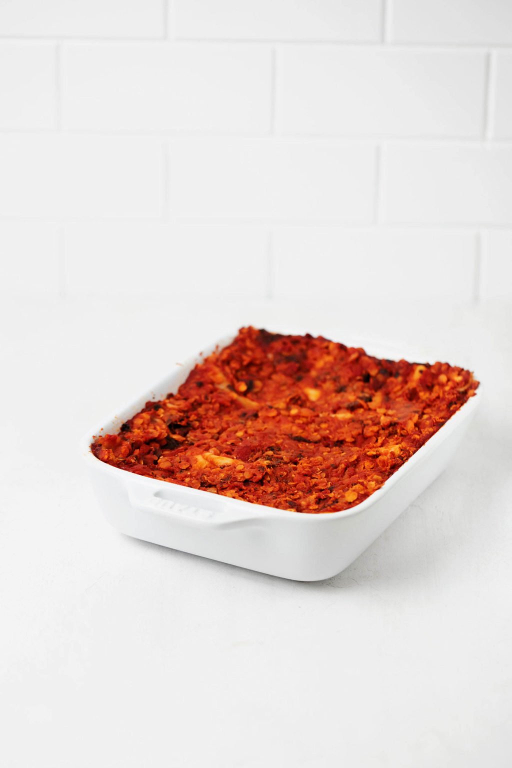 A dish of lasagna with a red, lentil-based sauce is resting on a white surface.