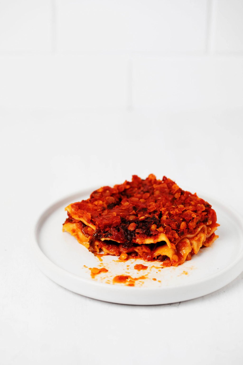 A square slice of lasagna rests on a small, round white plate, on top of a white surface.