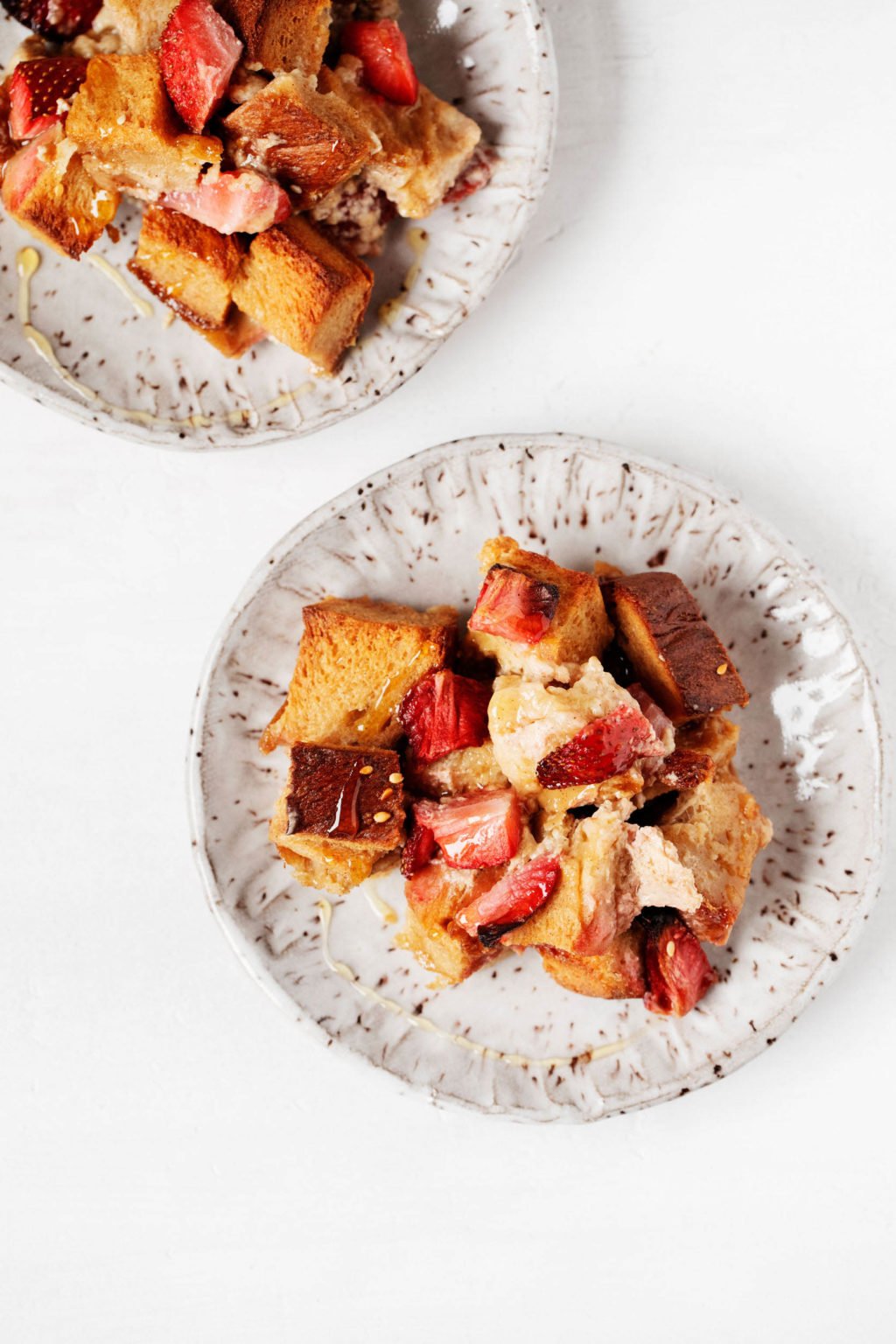 Two small, speckled white ceramic plates hold a vegan strawberry french toast casserole, made with cubed bread. 