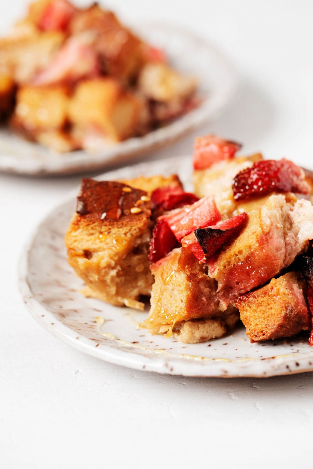 An angled photograph of two fluted dessert plates, which serve two portions of a baked French toast breakfast with strawberries. They rest on a white backdrop.