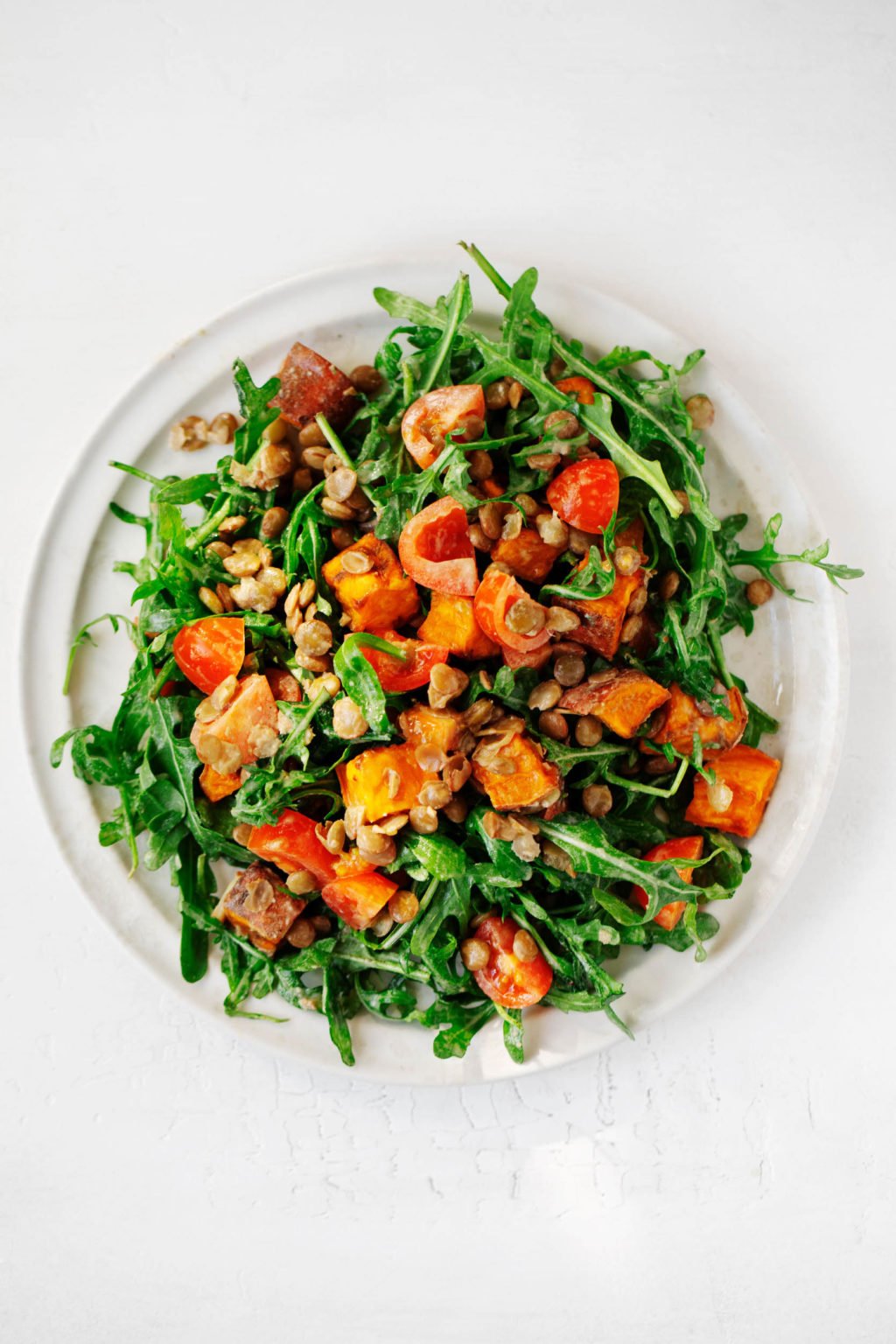 A round, white salad plate has been piled with arugula, sweet potatoes, grape tomatoes, lentils, and a creamy dressing.