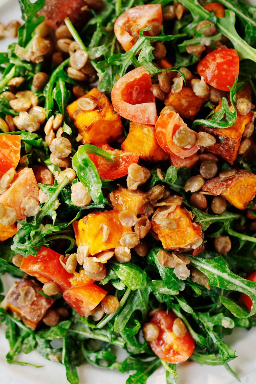 A close up image of a mixture of salad greens, roasted sweet potatoes, grape tomatoes, and lentils.