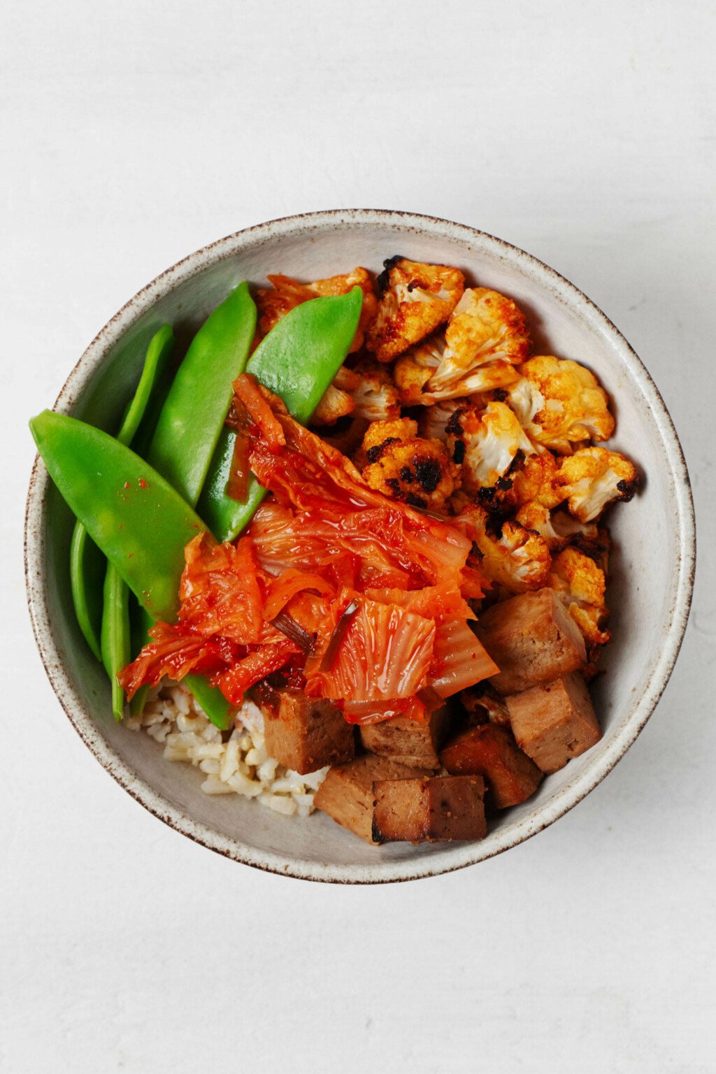 An overhead image of a plant-based bowl meal filled with kimchi, snow peas, tofu cubes, and cauliflower.