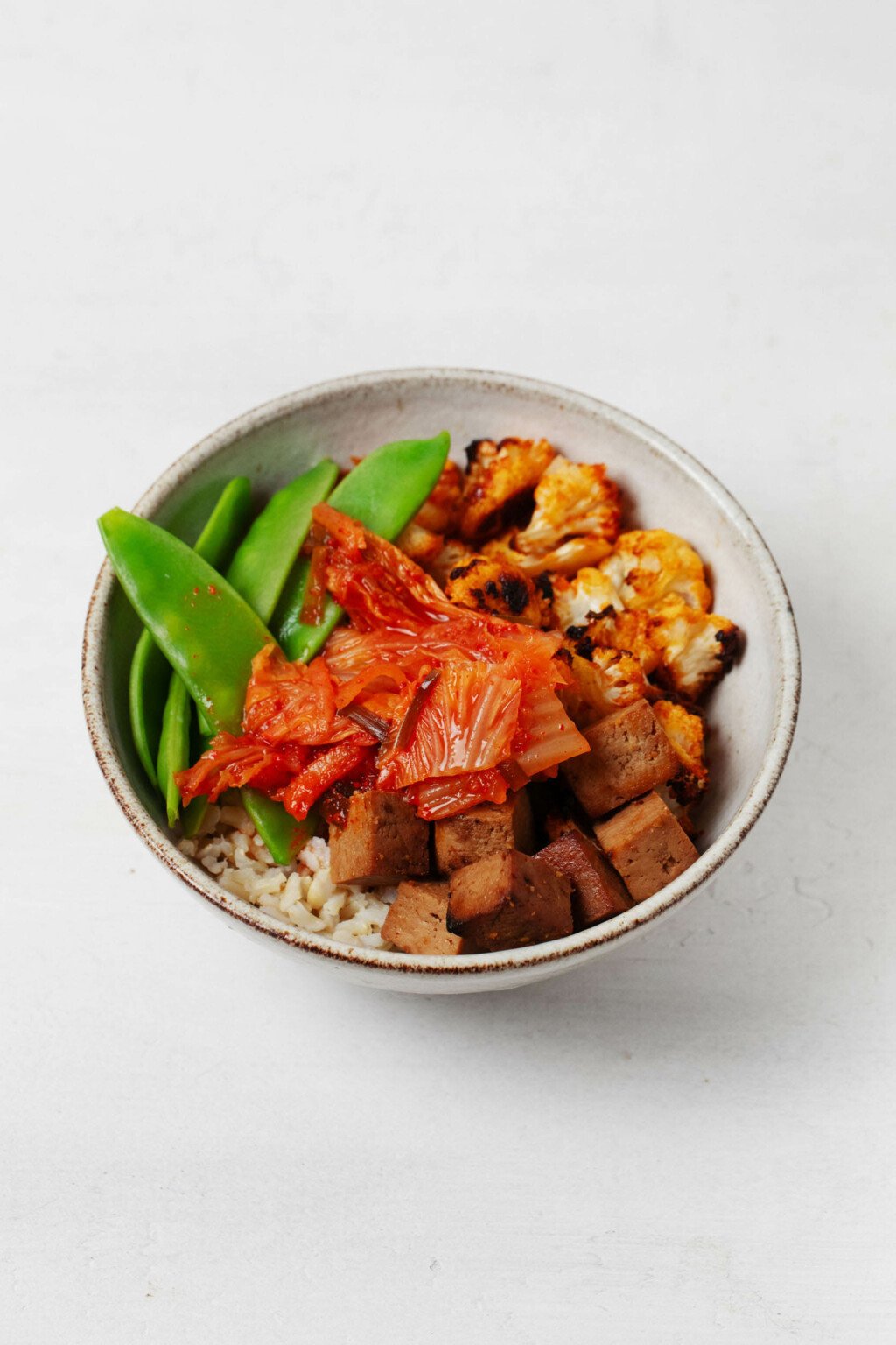 A white, ceramic bowl is resting on a white surface. It's filled with colorful, vegan vegetables and whole grains.