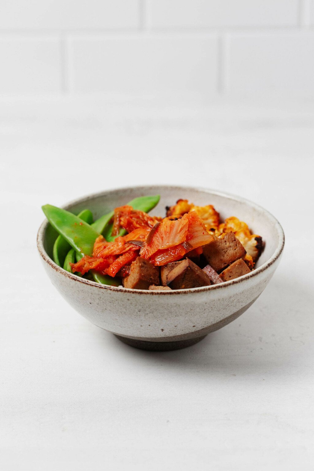 A round, white ceramic bowl holds a mixture of plant-based ingredients. It rests on a white surface.