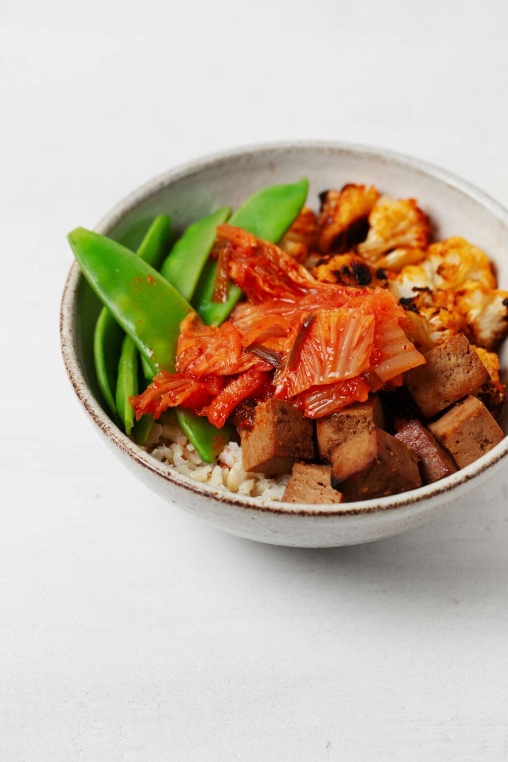 A chili roasted cauliflower and brown rice kimchi bowl is resting on a white surface.