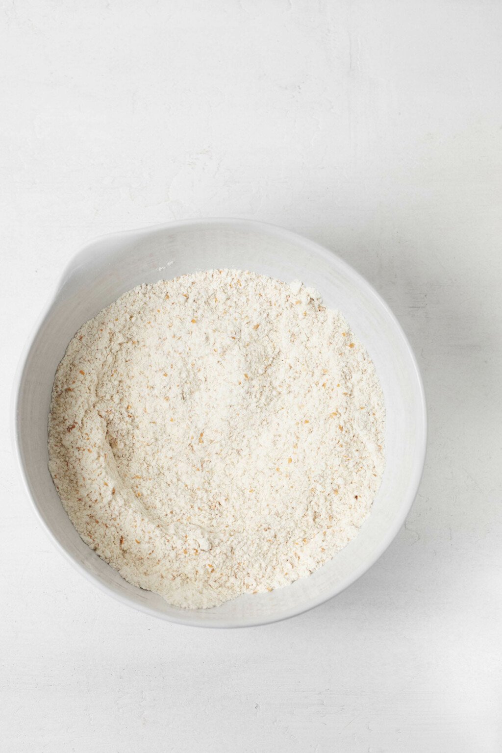 A white mixing bowl is filled with flour. It rests on a white surface.