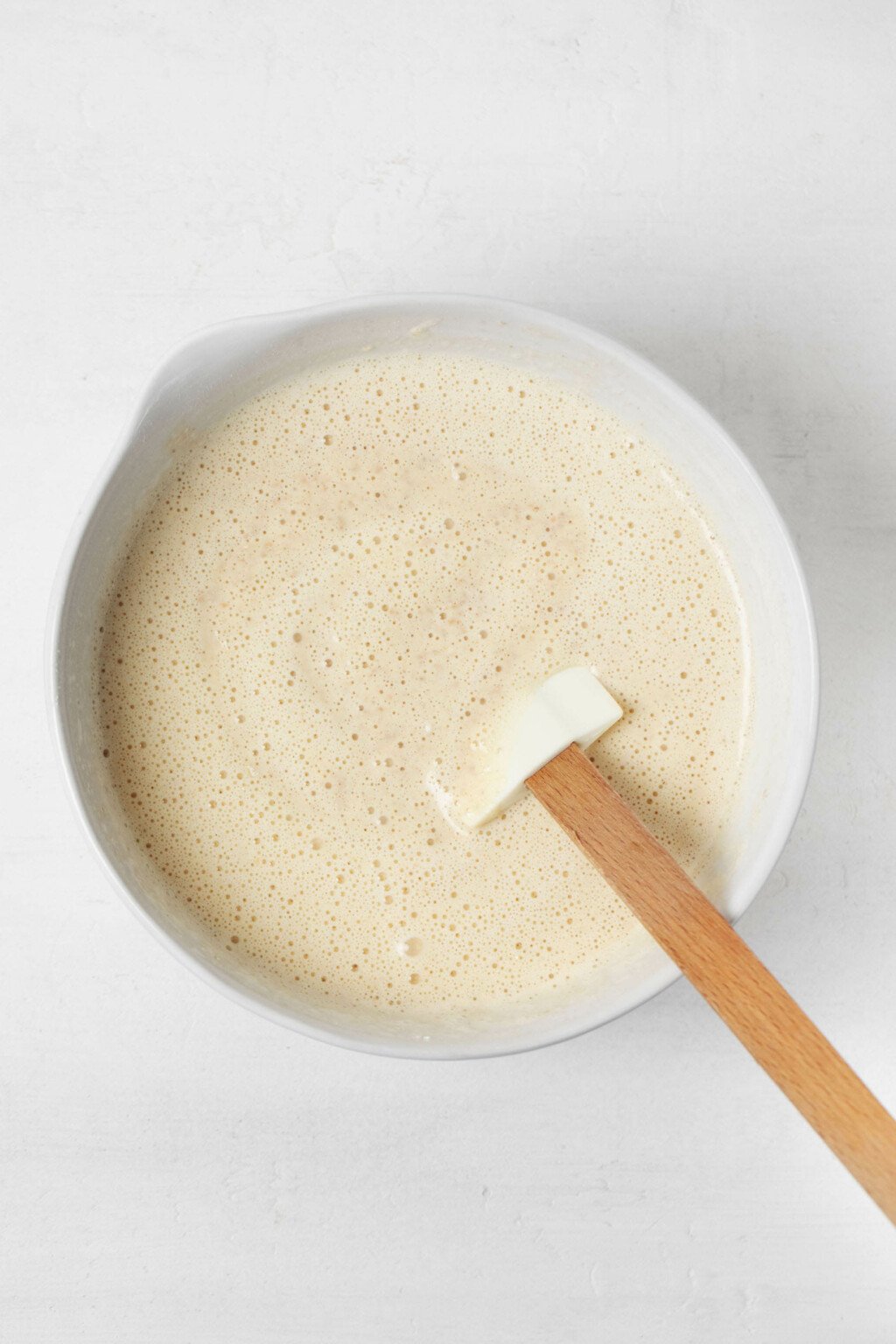 A white mixing bowl is filled with waffle batter. A spatula with a wooden handle pokes out of the bowl.