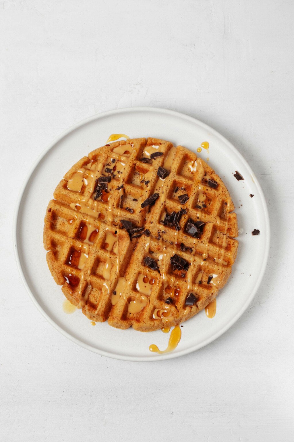 An overhead image of a round, white plate, which is holding a light and crispy vegan peanut butter waffle. The waffle is topped with chopped dark chocolate and a drizzle of peanut butter.